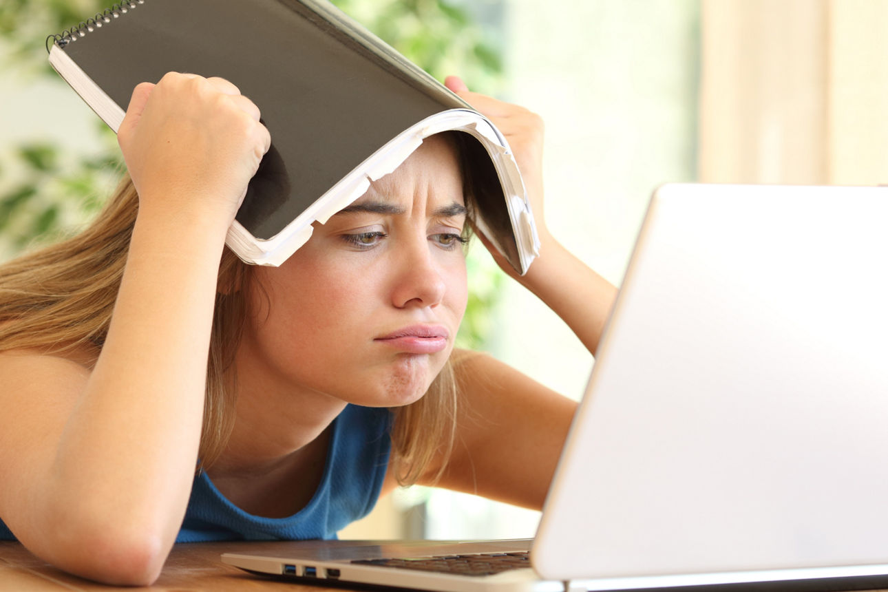 Mädchen schaut genervt in Laptop und hält sich die Hausaufgaben über den Kopf