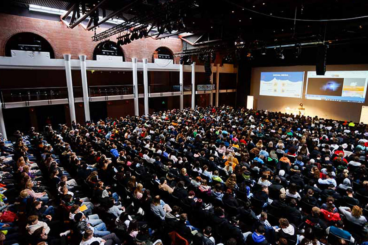Saal mit mehreren hundert Menschen und einer Bühne mit einer Präsentation