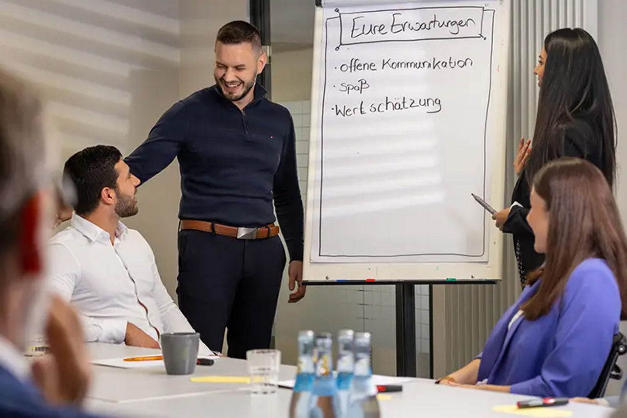 Mitarbeiter steht einem Flipchart und scherzt mit einem sitzendem Kollegen