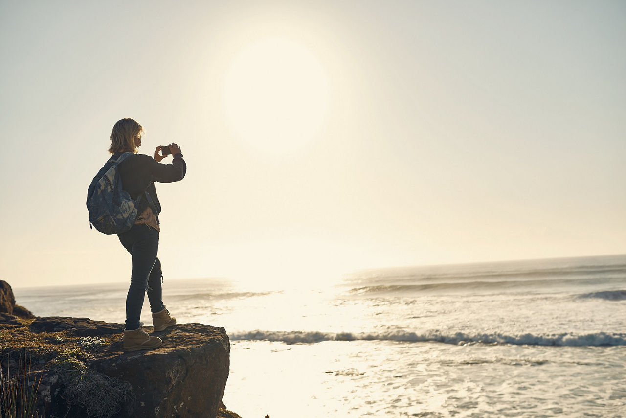 Eine Frau mit Rucksack steht an einer Klippe und macht ein Foto vom offenen Meer. 