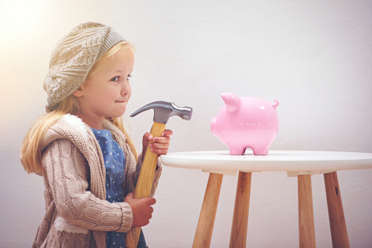 Kleines Mädchen mit Mütze zielt mit einem Hammer auf ein rosa Sparschwein