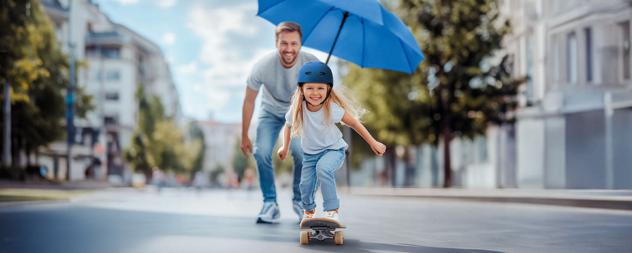 Mädchen fährt auf einem Skateboard, Vater mit blauem Schirm steht dahinter, im Hintergrund eine Straße und Häuser