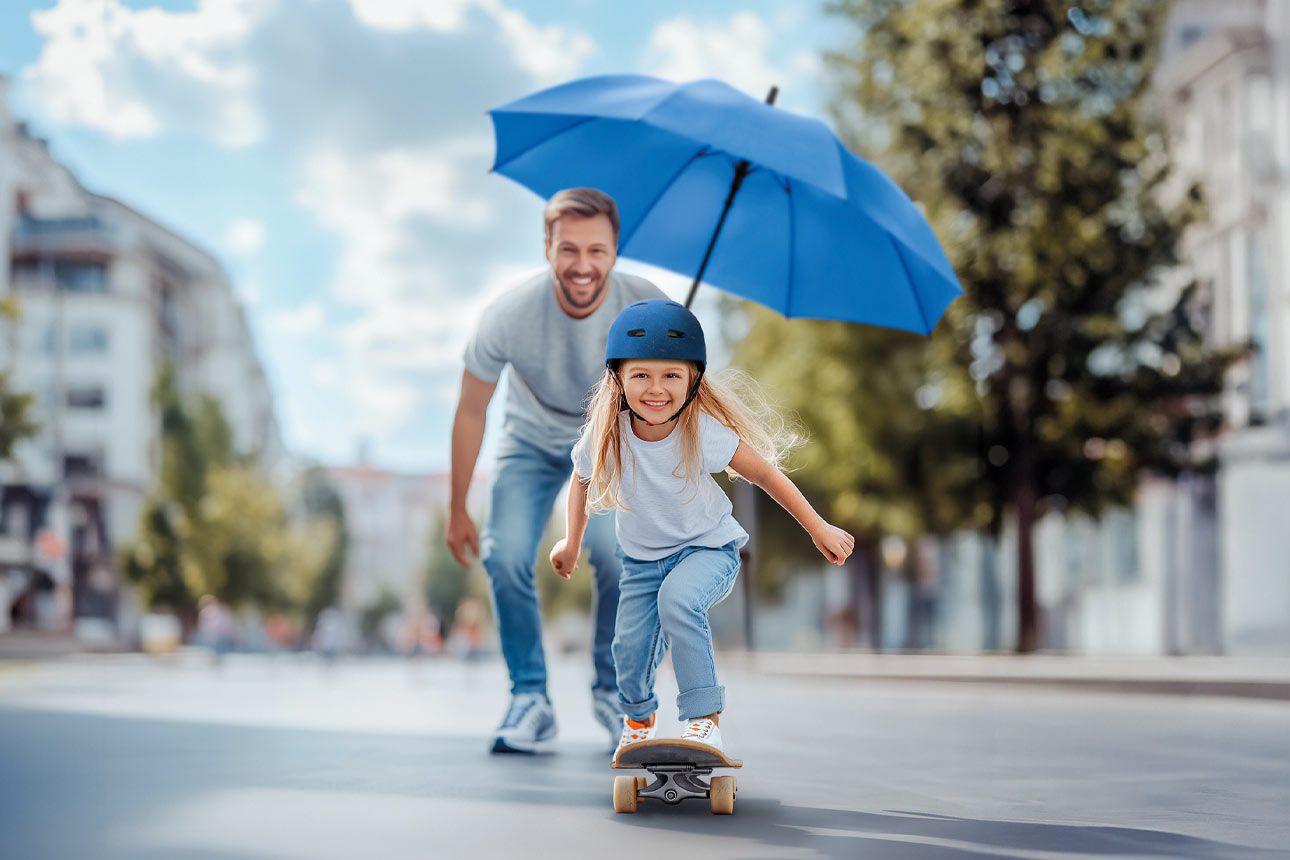 Mädchen fährt auf einem Skateboard, Vater mit blauem Schirm steht dahinter, im Hintergrund eine Straße und Häuser