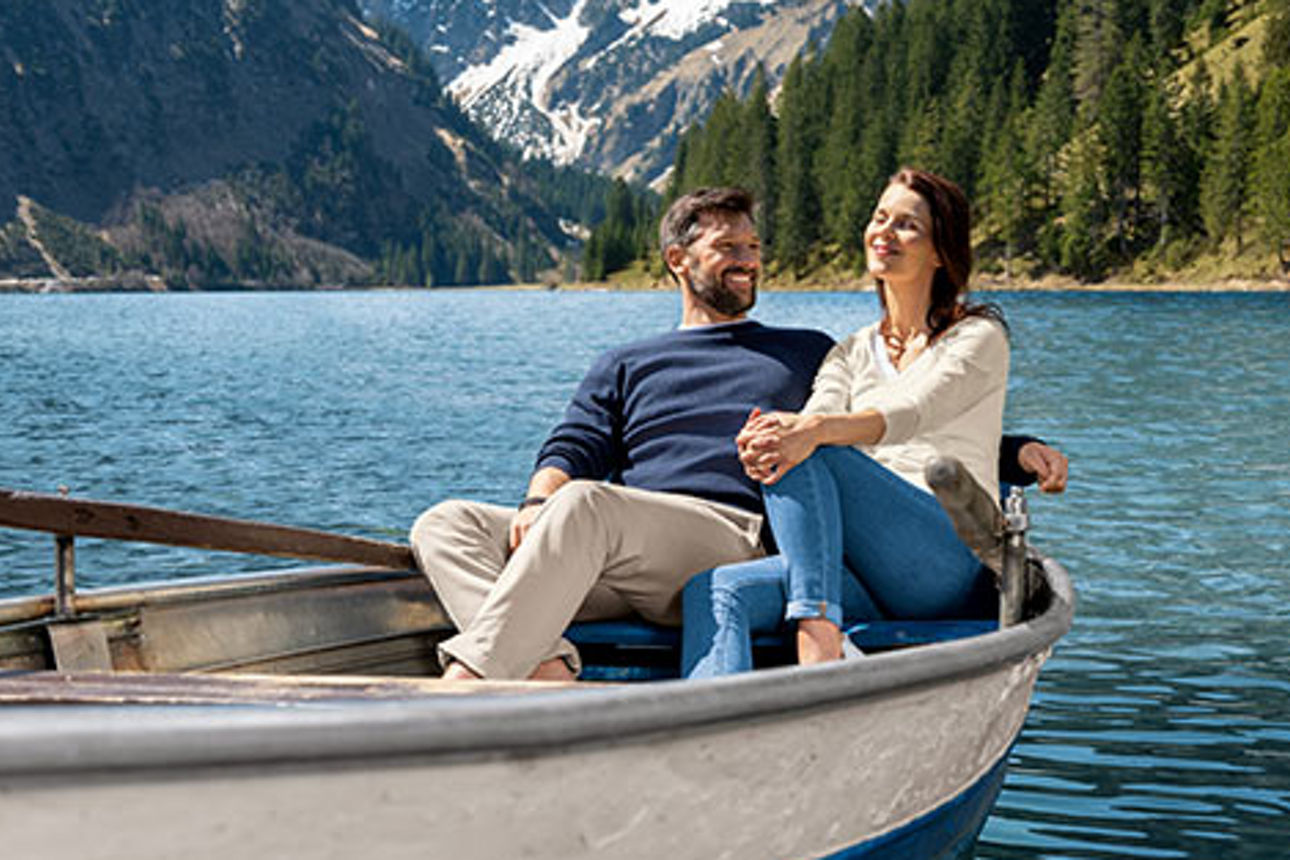 Mann und Frau sitzen entspannt in einem Paddelboot auf einem See, Berge im Hintergrund