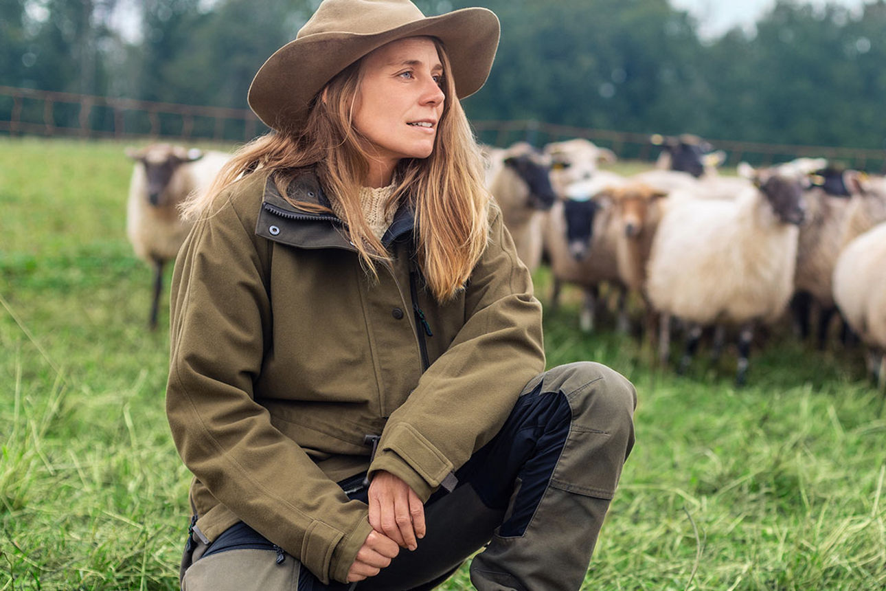 Junge Frau mit Hut hockt auf einer grünen Wiese, im Hintergrund eine Schafherde