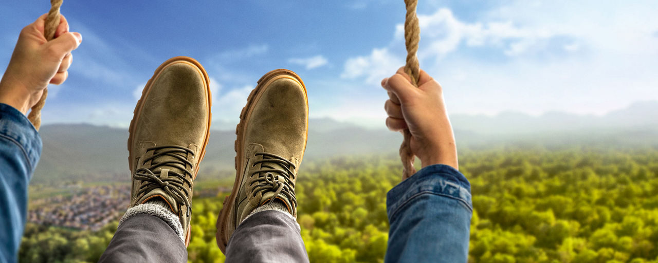 Die Hände und Füße einer Person auf einer Schaukel schwingen ins Bild, im Hintergrund Wald und Hügel