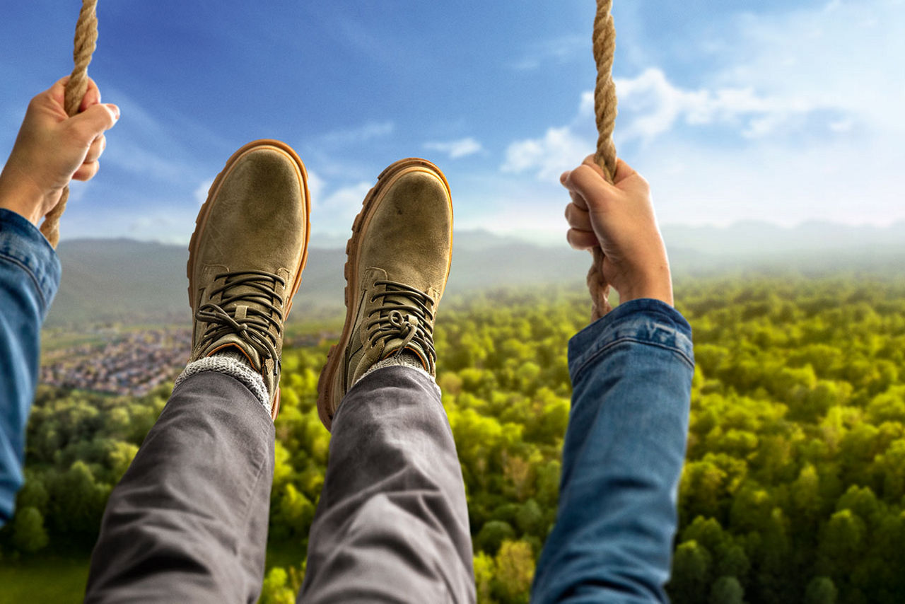 Die Hände und Füße einer Person auf einer Schaukel schwingen ins Bild, im Hintergrund Wald und Hügel