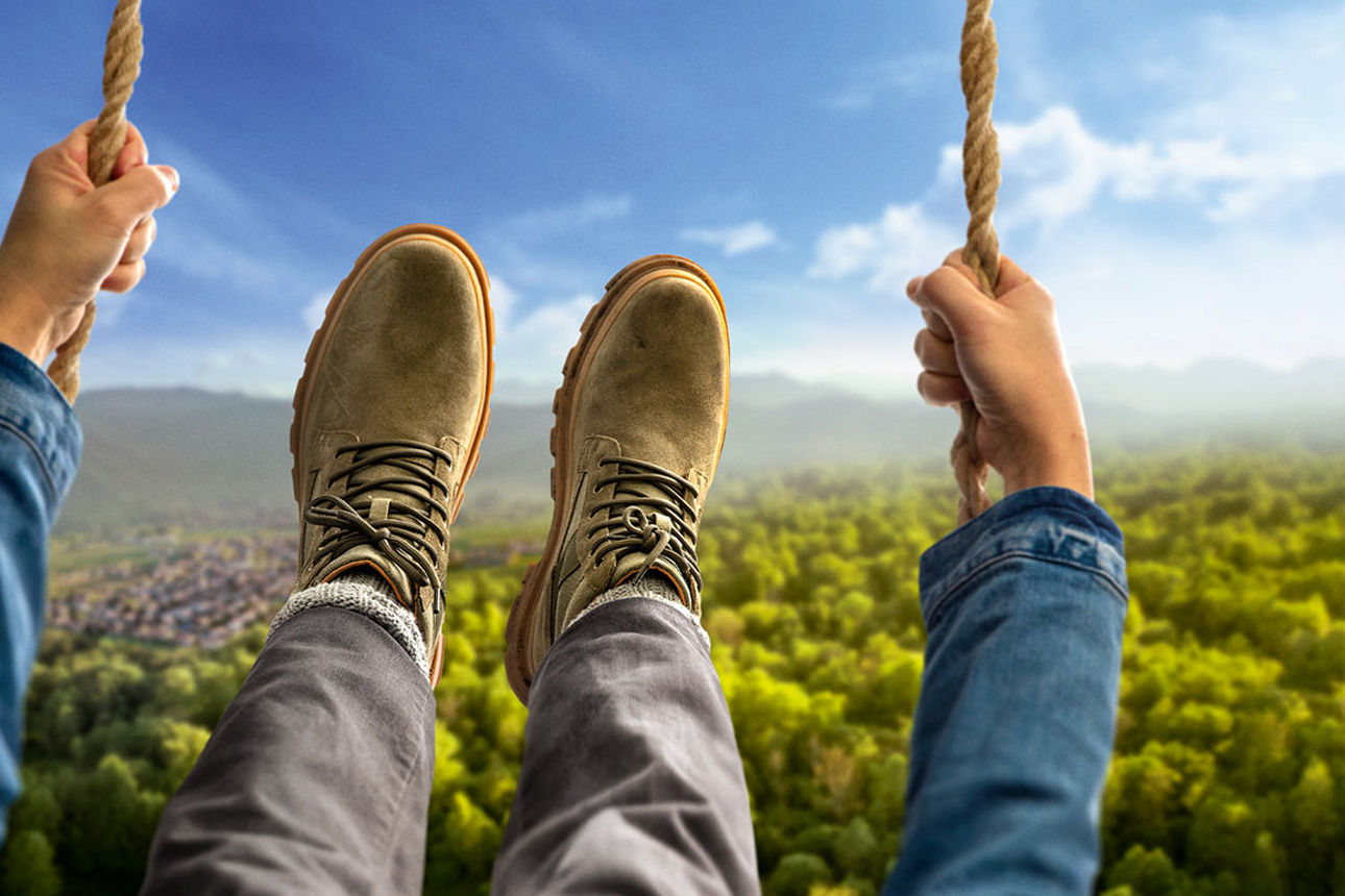 Die Hände und Füße einer Person auf einer Schaukel schwingen ins Bild, im Hintergrund Wald und Hügel