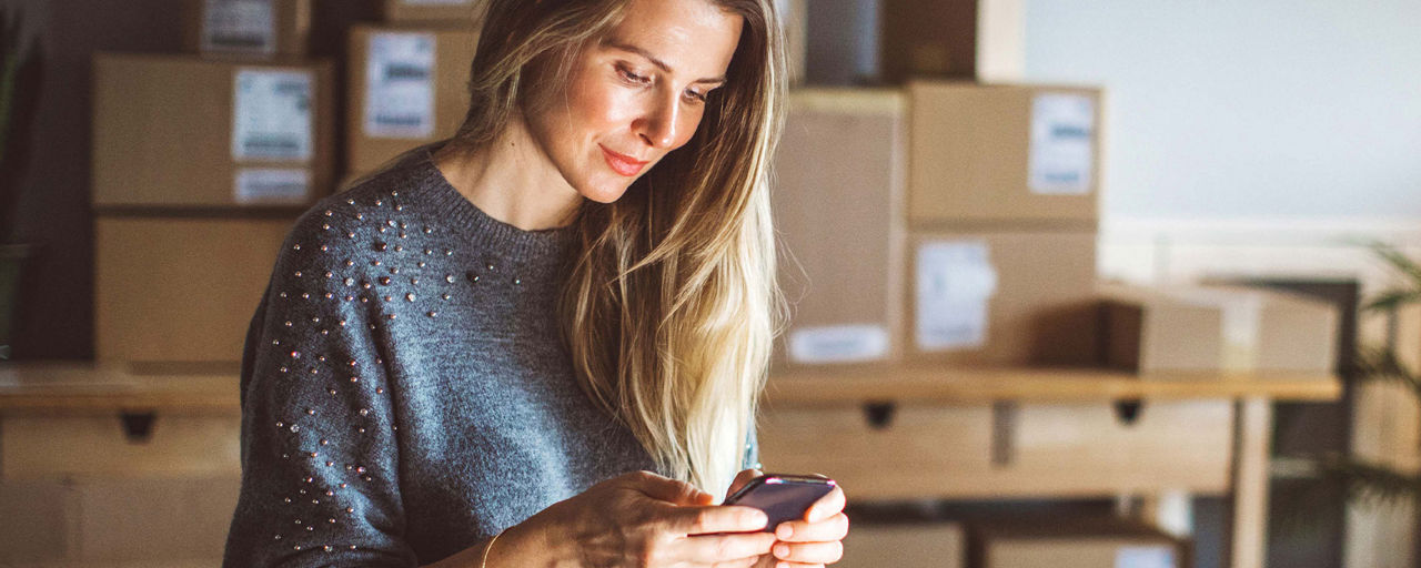 Eine Frau schaut auf ihr Smartphone; im Hintergrund Pakete