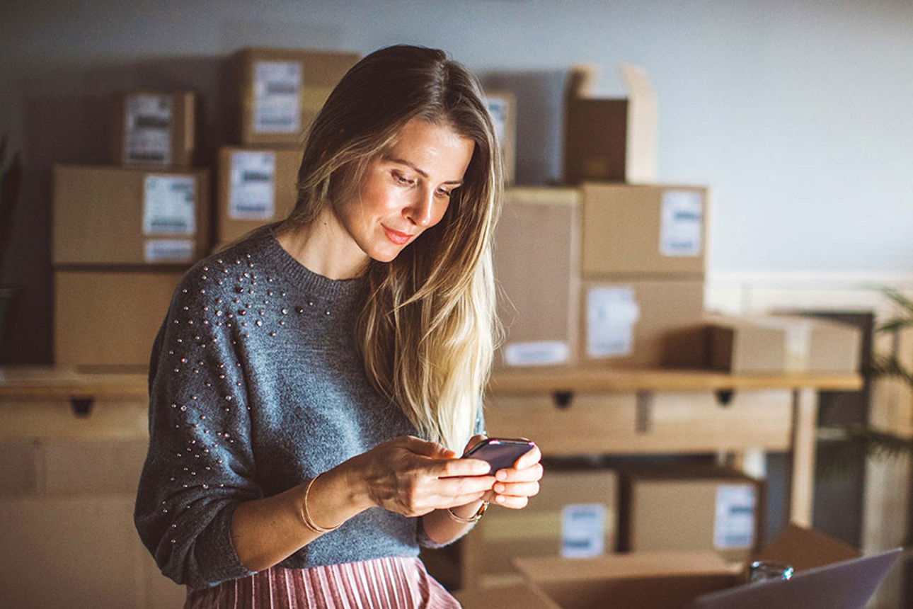 Frau schaut auf ihr Smartphone und im Hintergrund stehen aufgestappelt Kartons