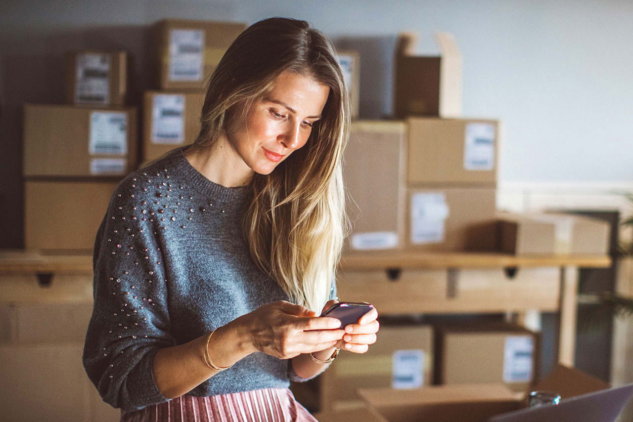 Eine Frau schaut auf ihr Smartphone; im Hintergrund Pakete