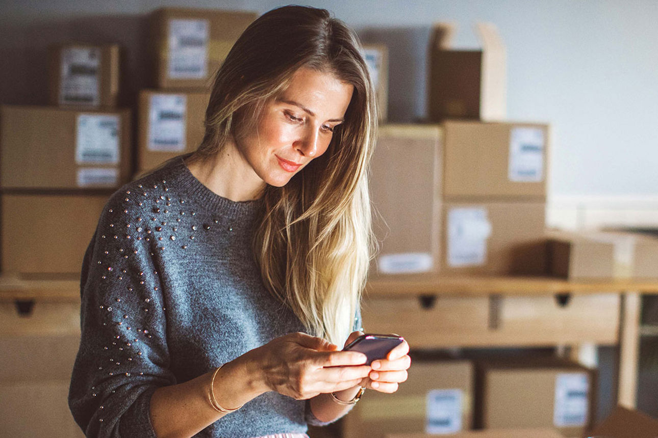 Eine Frau schaut auf ihr Smartphone; im Hintergrund Pakete