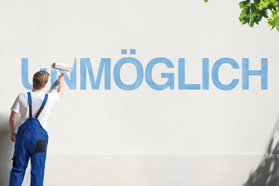 Junger Mann in Latzhose übermalt den Schriftzug "Unmöglich" auf einer Wand