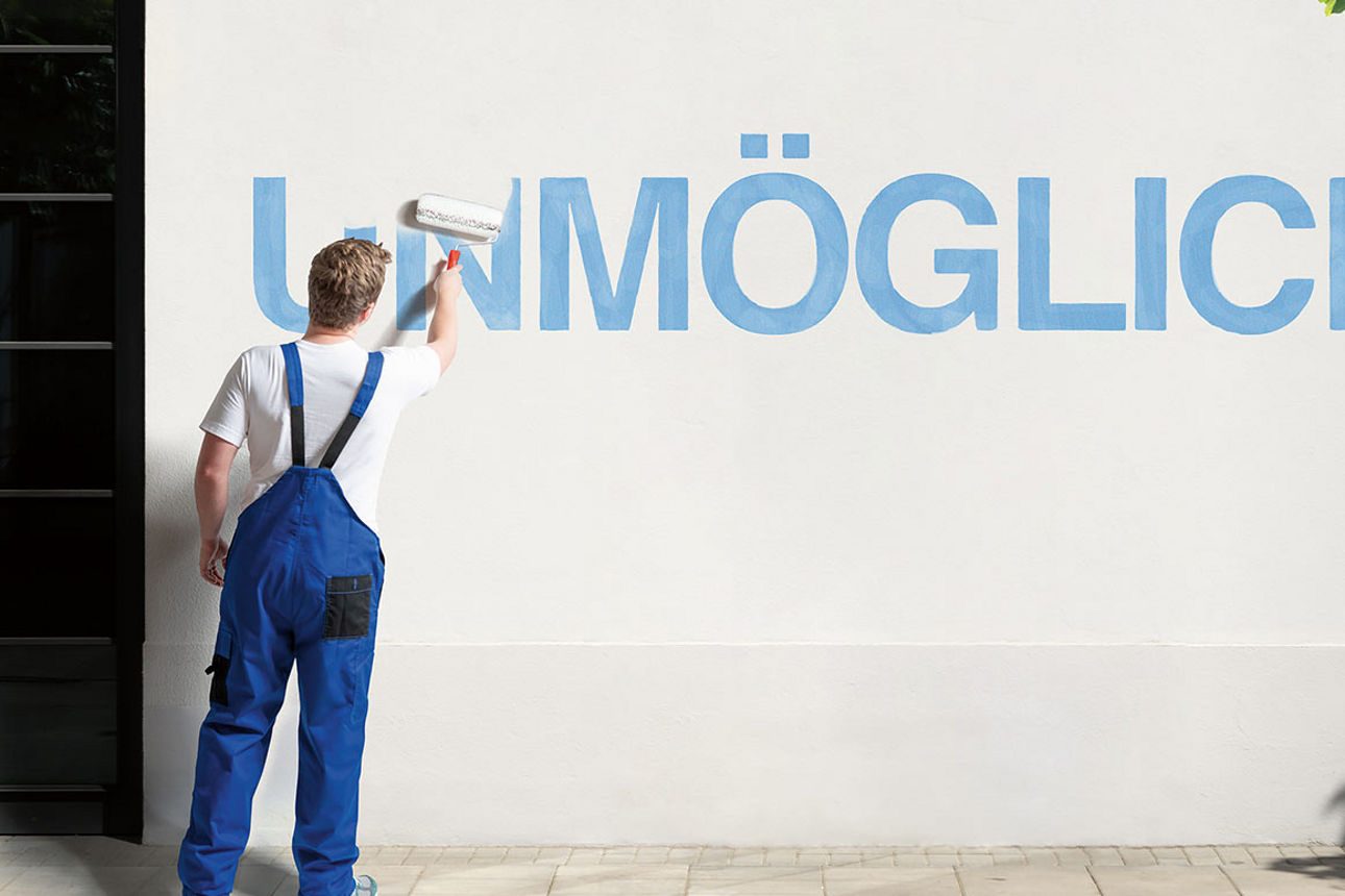 Junger Mann in Latzhose übermalt den Schriftzug „Unmöglich“ auf einer Wand