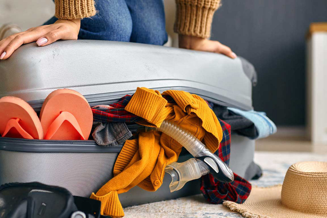 Eine Person kniet auf einem überfüllten, silbernen Koffer, aus dem Kleidung, Flip-Flops und eine Tauchmaske herausquellen