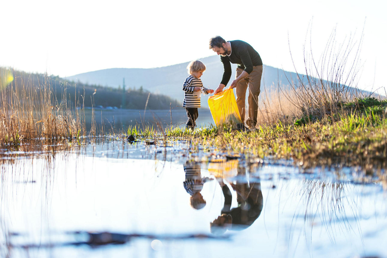Vater und stehen an einem Wasserlauf und sammeln Müll in eine Mülltüte
