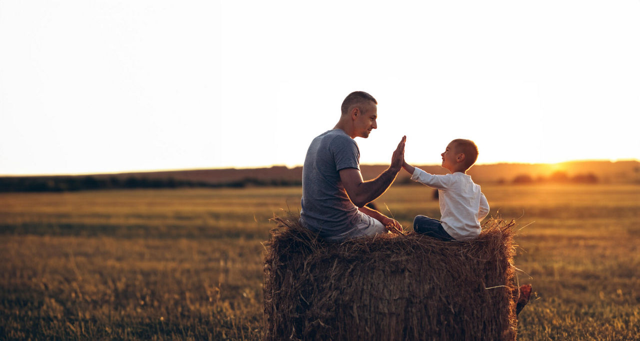 Vater und Sohn sitzen auf einem Stohballen bei Sonnenuntergang. 