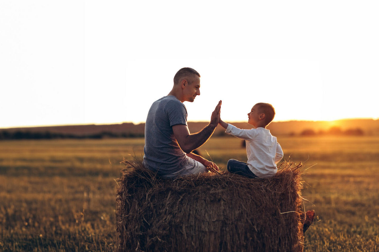 Vater und Sohn sitzen auf einem Stohballen bei Sonnenuntergang. 