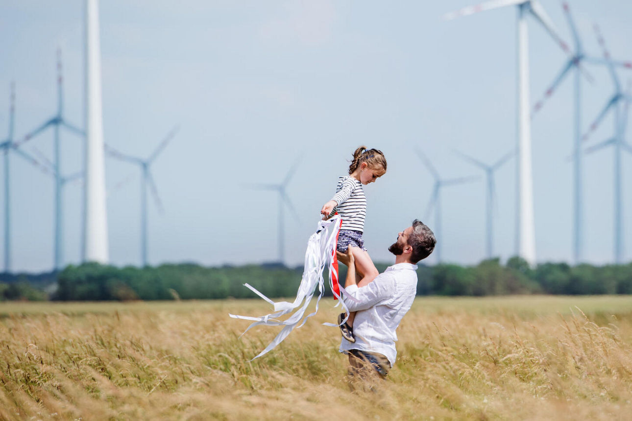 Vater hält seine Tochter vor sich hoch. Sie stehen auf einem Feld mit Windädern im Hintergrund.