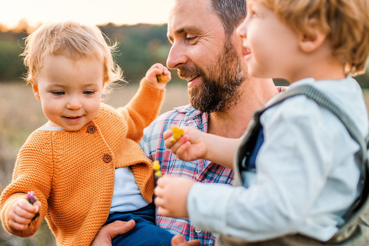 Ein Mann steht mit zwei kleinen Kindern auf dem Arm auf einer Wiese.