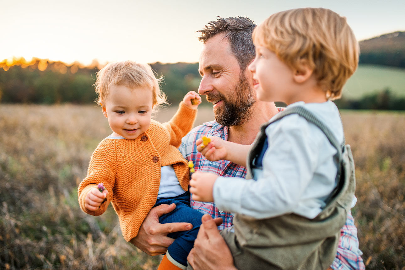 Vater mit zwei Kindern auf dem Arm steht auf einer Wiese