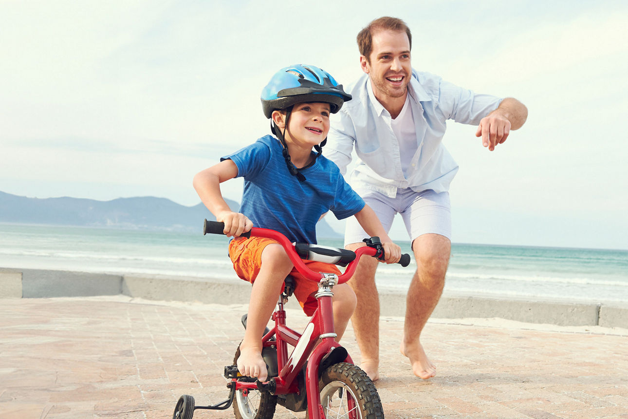 Kind auf Fahrrad mit Stützrädern, wird auf einem gepflasterten Weg am Strand vom Vater angeschoben.