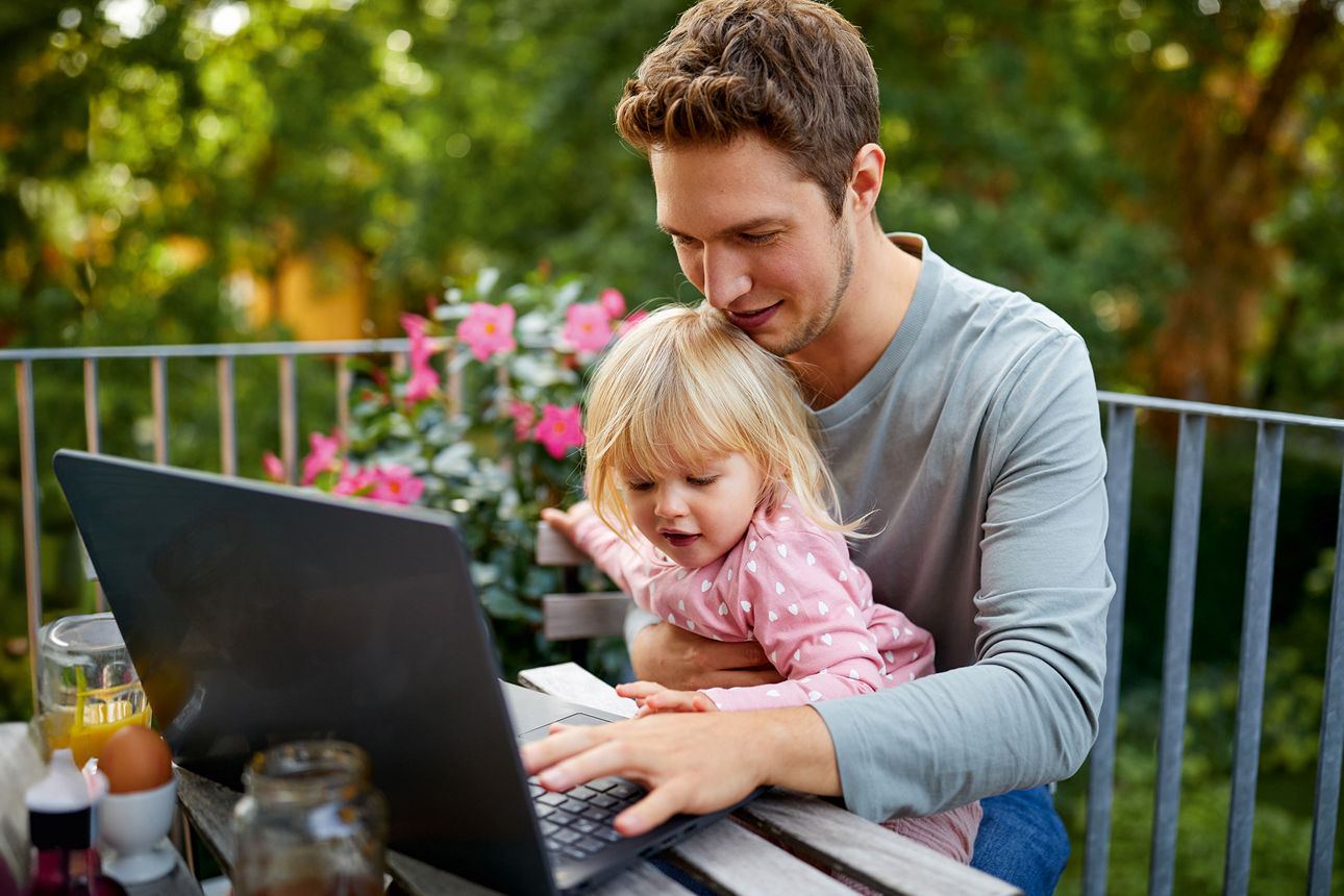Vater sitzt mit Kleinkind auf dem Schoß auf einem Balkon vor dem Laptop.