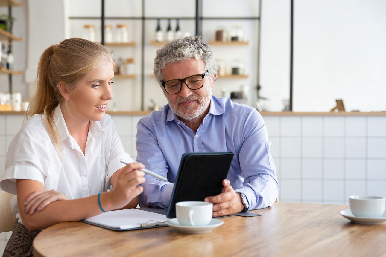 Junge Frau und Mann mit grauen Haaren sitzen zusammen am Tisch und schauen in einen Laptop