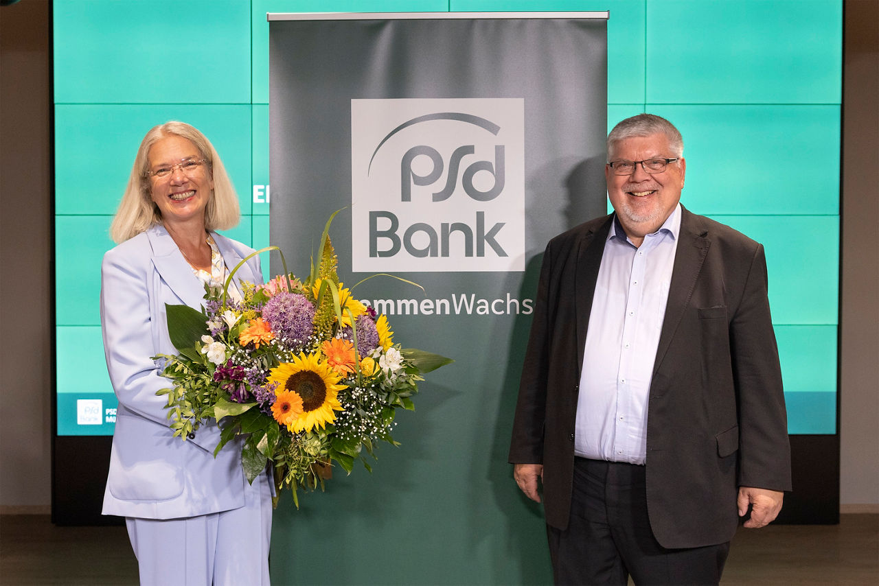 Eine Frau im hellblauen Anzug hält einen großen Blumenstrauß in der Hand.  Rechts im Bild steht ein Mann im Anzug. In der Mitte sieht man ein grünes Banner der PSD Bank. 