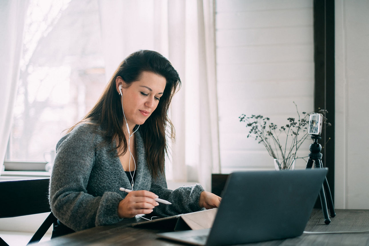 Frau sitzt an einem Tisch, hat Kopfhörer auf und notiert etwas auf einem Tablet. Vor ihr steht ein aufgeklappter Laptop