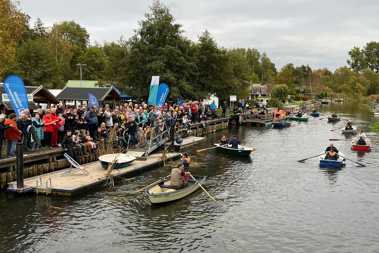 Auf einem Fluß (die Recknitz) rudern ein paar Ruderboote. Am Ufer steht eine Menschenmenge und Fahnen vom NDR und der Volksbank sind zu sehen.