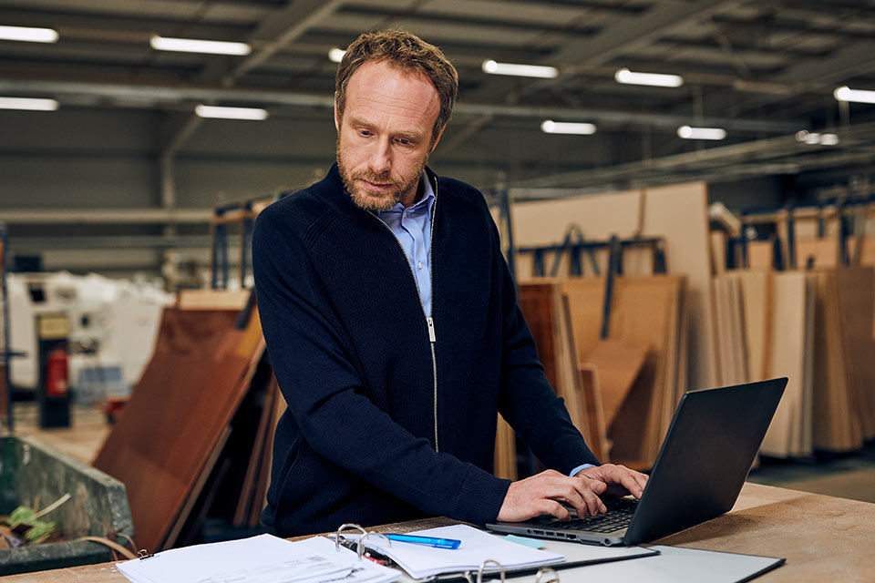 Mann steht in Schreinerei am Tisch und arbeitet am Laptop