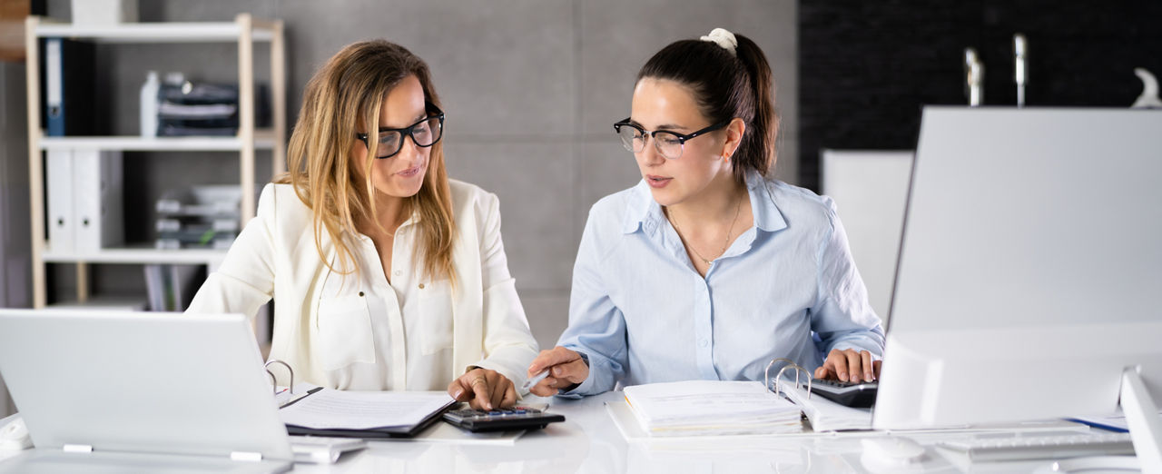 zwei Frauen in Hemd gucken auf Taschenrechner am Arbeitsplatz