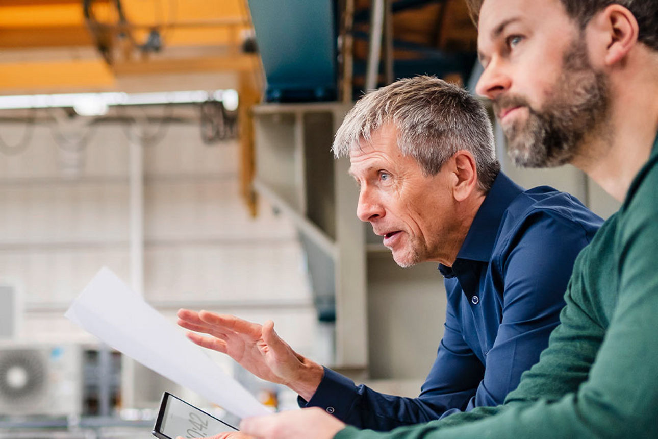 Zwei Männer stehen in Produktionshalle, einer der Männer hält ein Blatt in der Hand.
