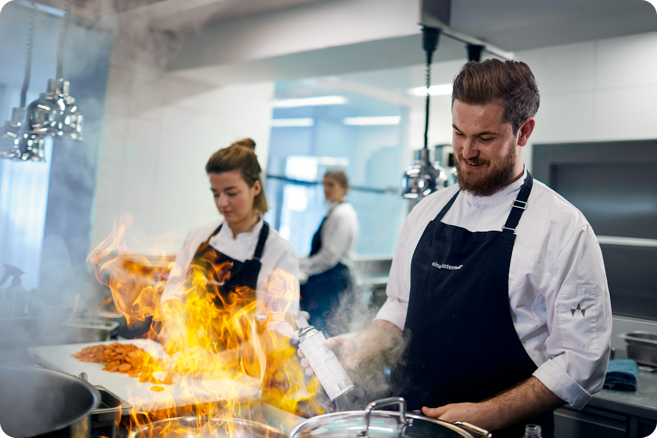 Ein Koch und eine Köchin stehen in der Küche und flambieren ein Gericht