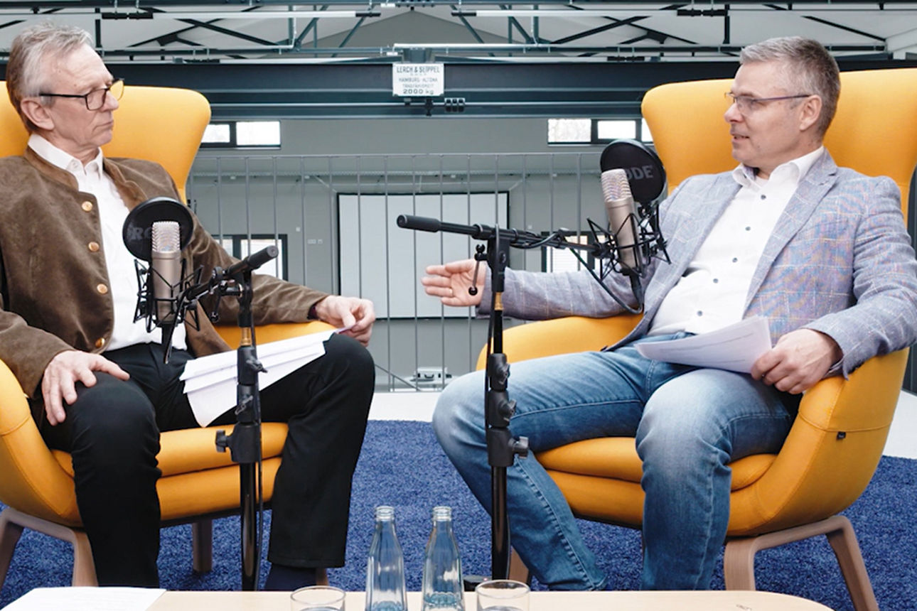 Zwei Personen im Gespräch vor Mikrofonen, Podcast- oder Interviewaufnahme in modernem Studio.