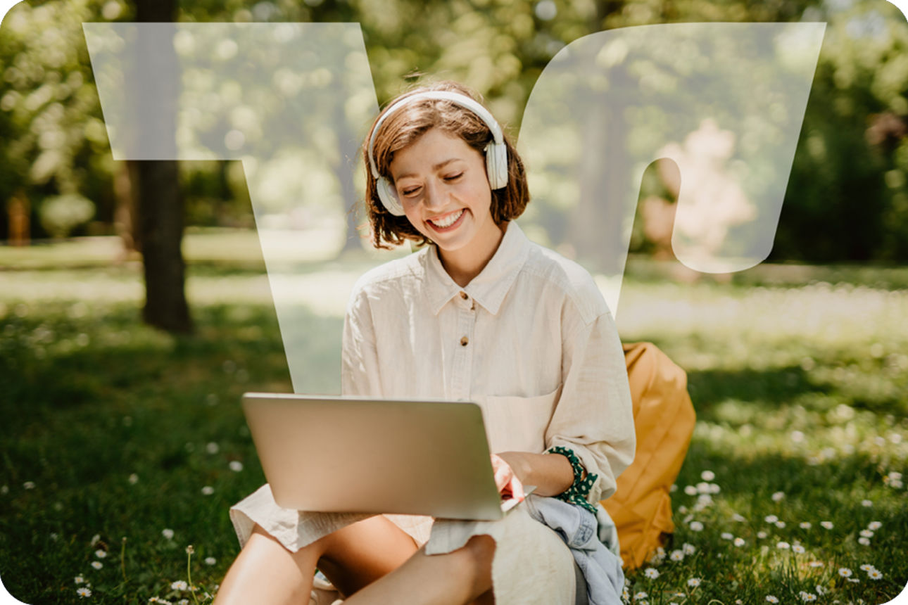 Person sitzt im Park auf dem Gras, trägt Kopfhörer und arbeitet an einem Laptop.
