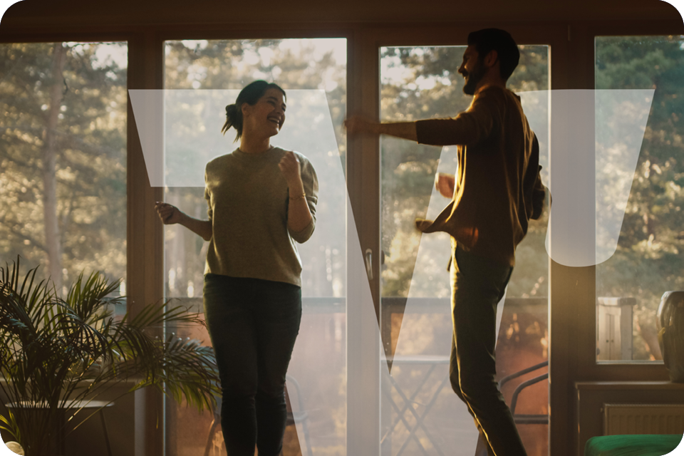 Zwei Personen tanzen vor großen Fensterfronten in einem lichtdurchfluteten Wohnzimmer mit Blick ins Grüne.