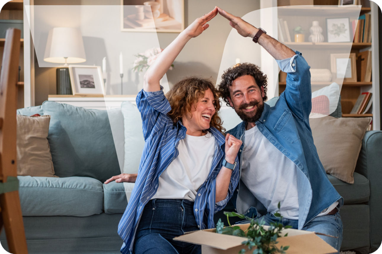 Ehepaar sitzt im Wohnzimmer auf dem Boden, formen gemeinsam mit den Armen ein Dach über sich 