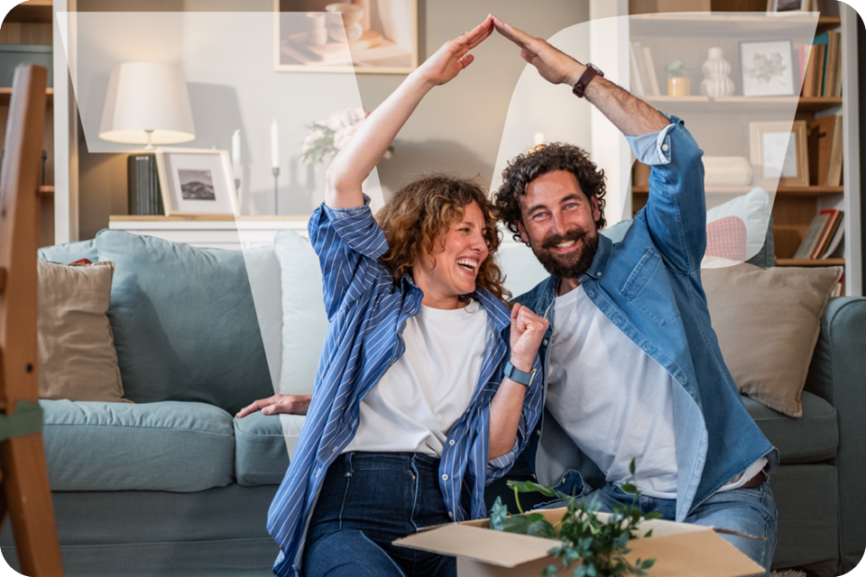Ehepaar sitzt im Wohnzimmer auf dem Boden, formen gemeinsam mit den Armen ein Dach über sich 