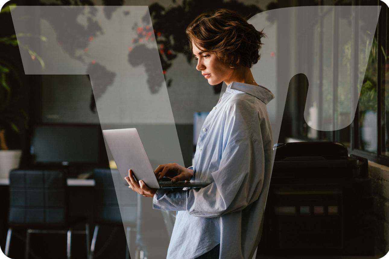 Person steht in einem modernen Büro und arbeitet an einem Laptop vor einer Weltkarte an der Wand.