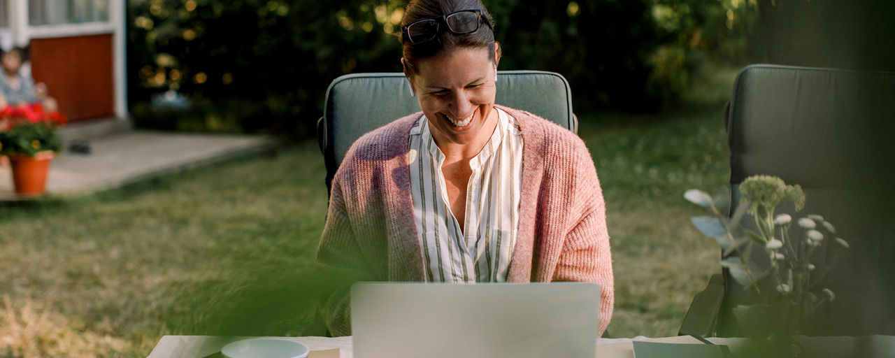 Eine Frau sitzt im Garten lachend vor ihrem Laptop
