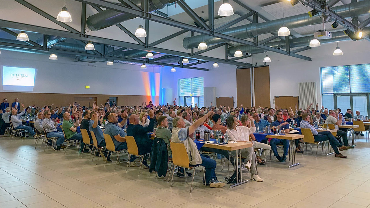 Mitglieder der Vertreterversammlung sitzen in der Wiedparkhalle an langen Tischreihen und heben die Hand zum Abstimmen