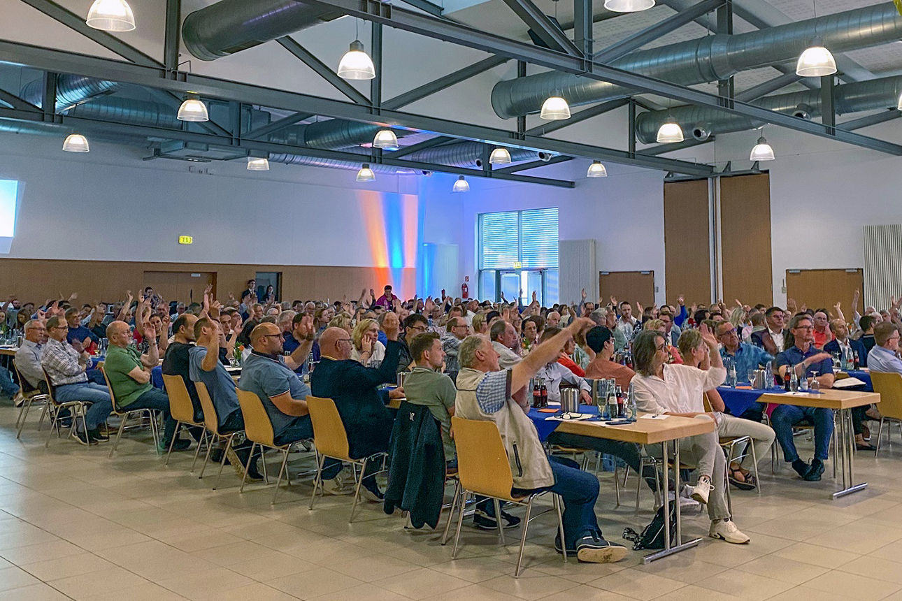 Mitgliedervertreterinnen und Vertreter sitzen in der Wiedparkhalle an langen Tischen und heben die Hand zur Abstimmung