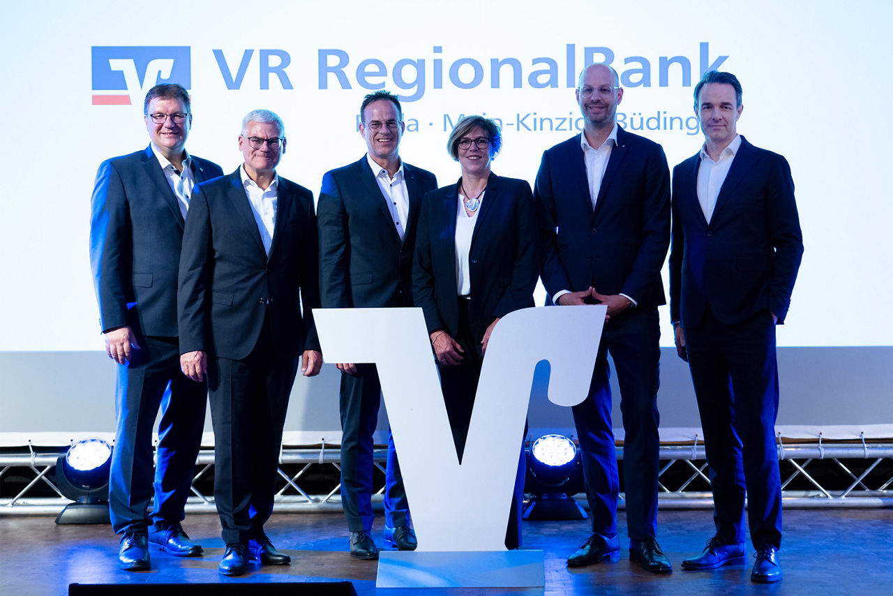 Das Bild zeigt die Vorstände der VR Bank Main-Kinzig-Büdingen und VR Bank Fulda,  vor einer Wand auf der das Logo der neuen VR RegionalBank zu sehen ist. 