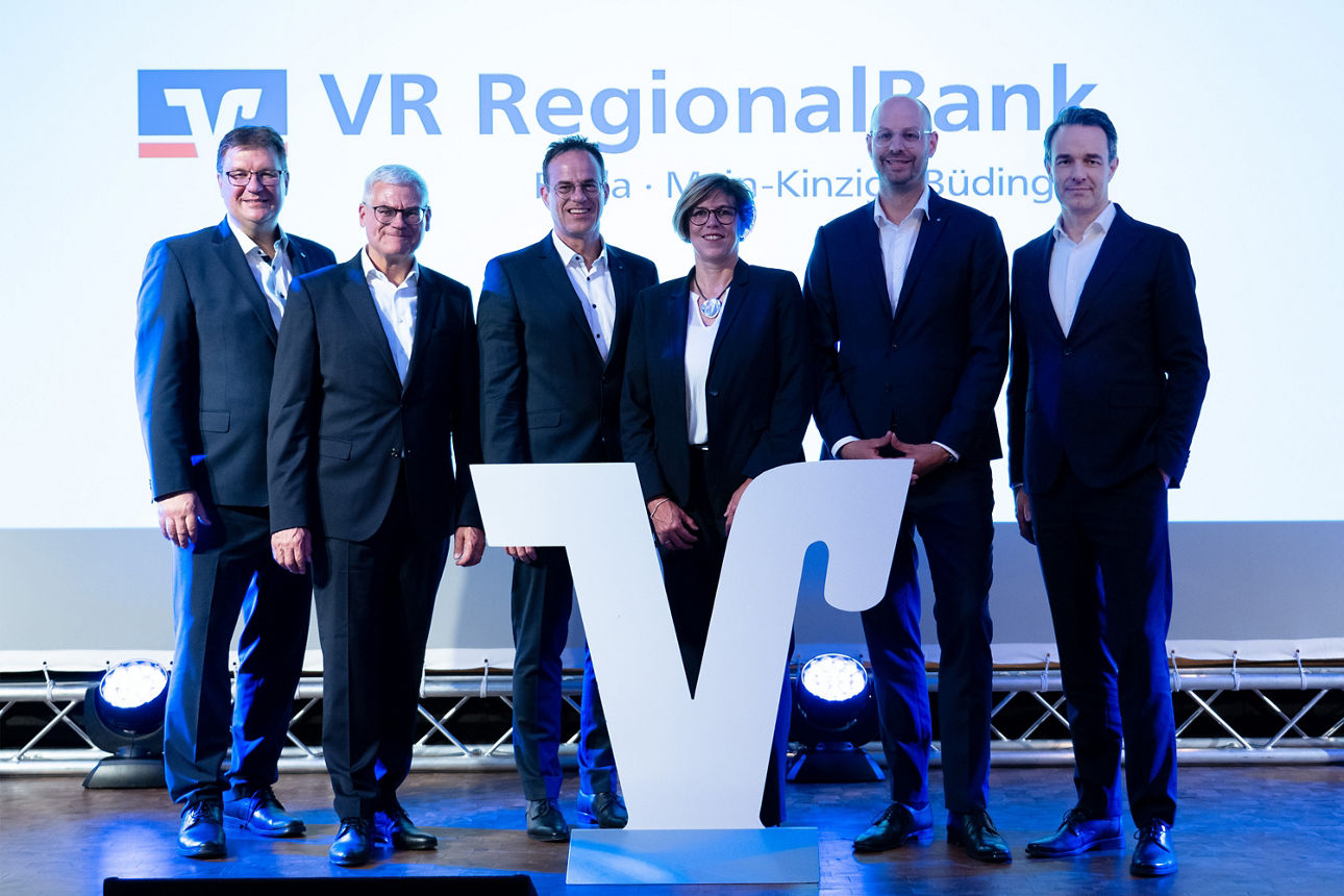 Das Bild zeigt die Vorstände der VR Bank Main-Kinzig-Büdingen und VR Bank Fulda,  vor einer Wand auf der das Logo der neuen VR RegionalBank zu sehen ist. 