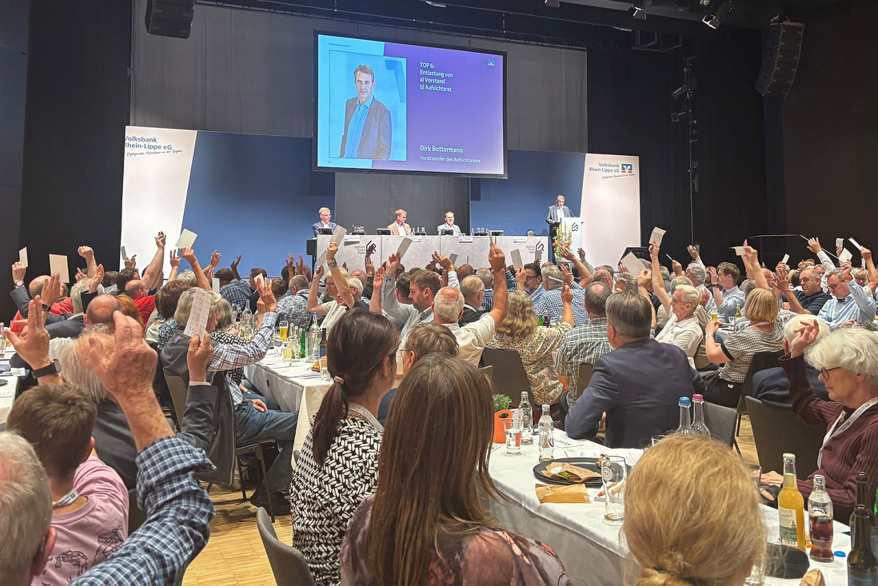 Eine große Gruppe von Personen, die in einer geräumigen Halle an Tischen sitzt und sich unterhält und beschäftigt.