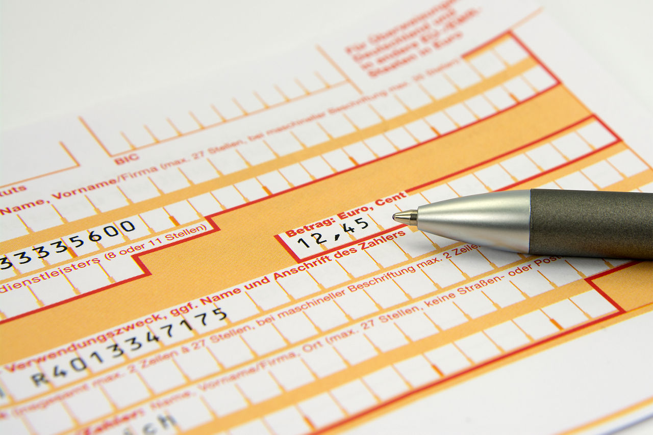 Das Bild zeigt einen ausgefüllten Überweisungsschein und einen Kugelschreiber.