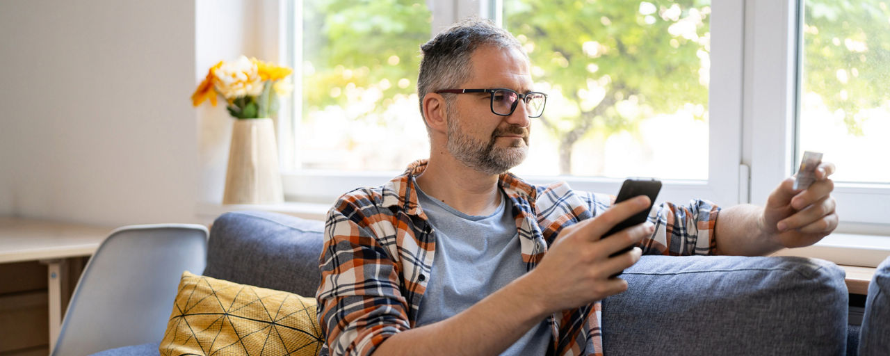 Ein Mann sitzt auf der Couch mit Smartphone und Personalausweis in der Hand