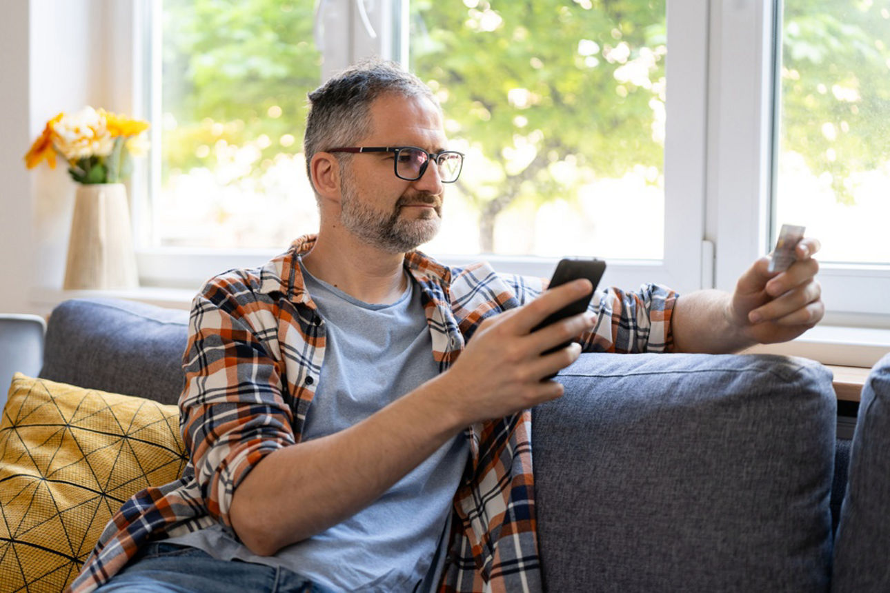 Ein Mann sitzt auf der Couch mit Smartphone und Personalausweis in der Hand